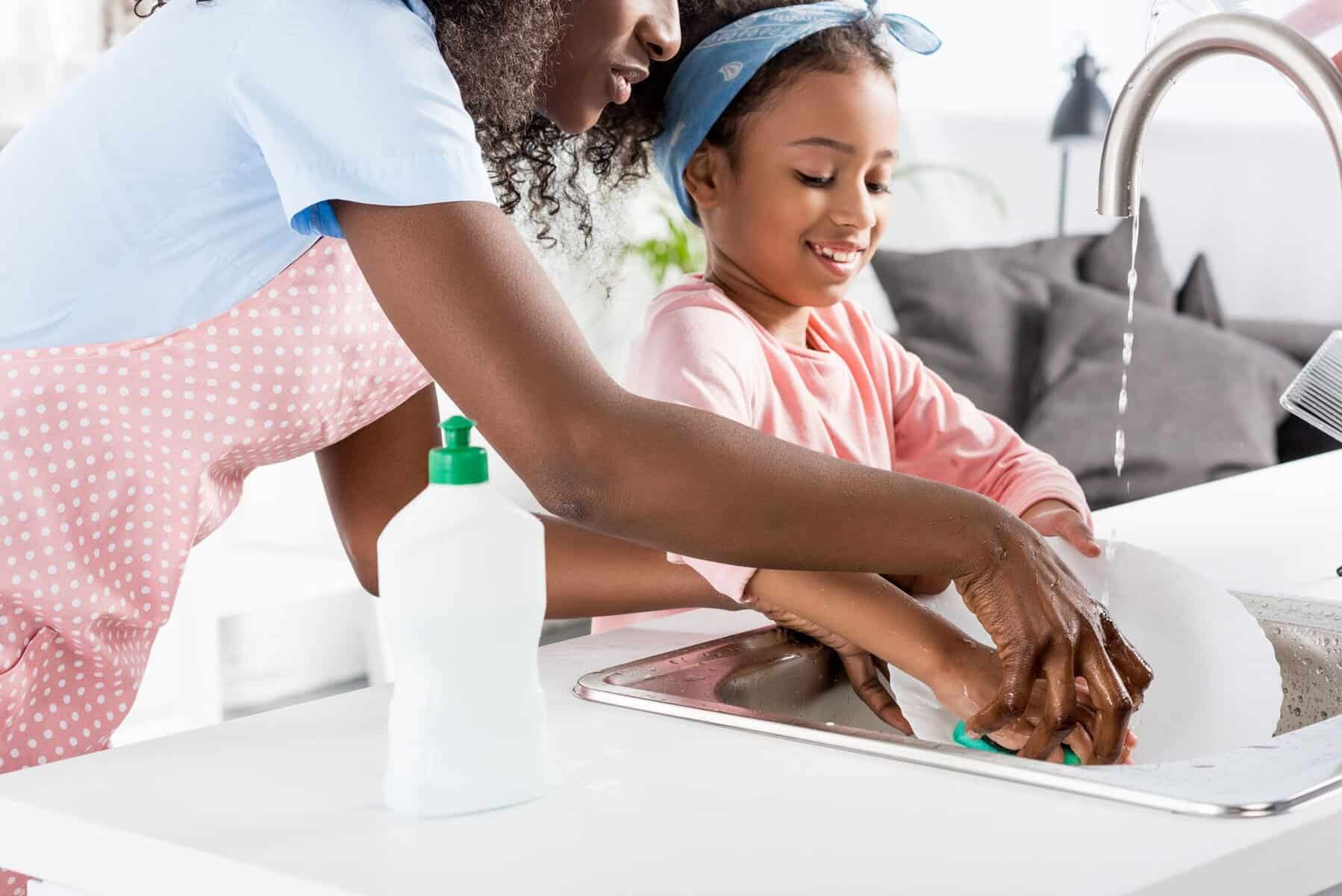 An adult and a child wash dishes together at a kitchen sink, with a soap bottle and running faucet visible—showing the comfort of choosing the best water heater for home use.