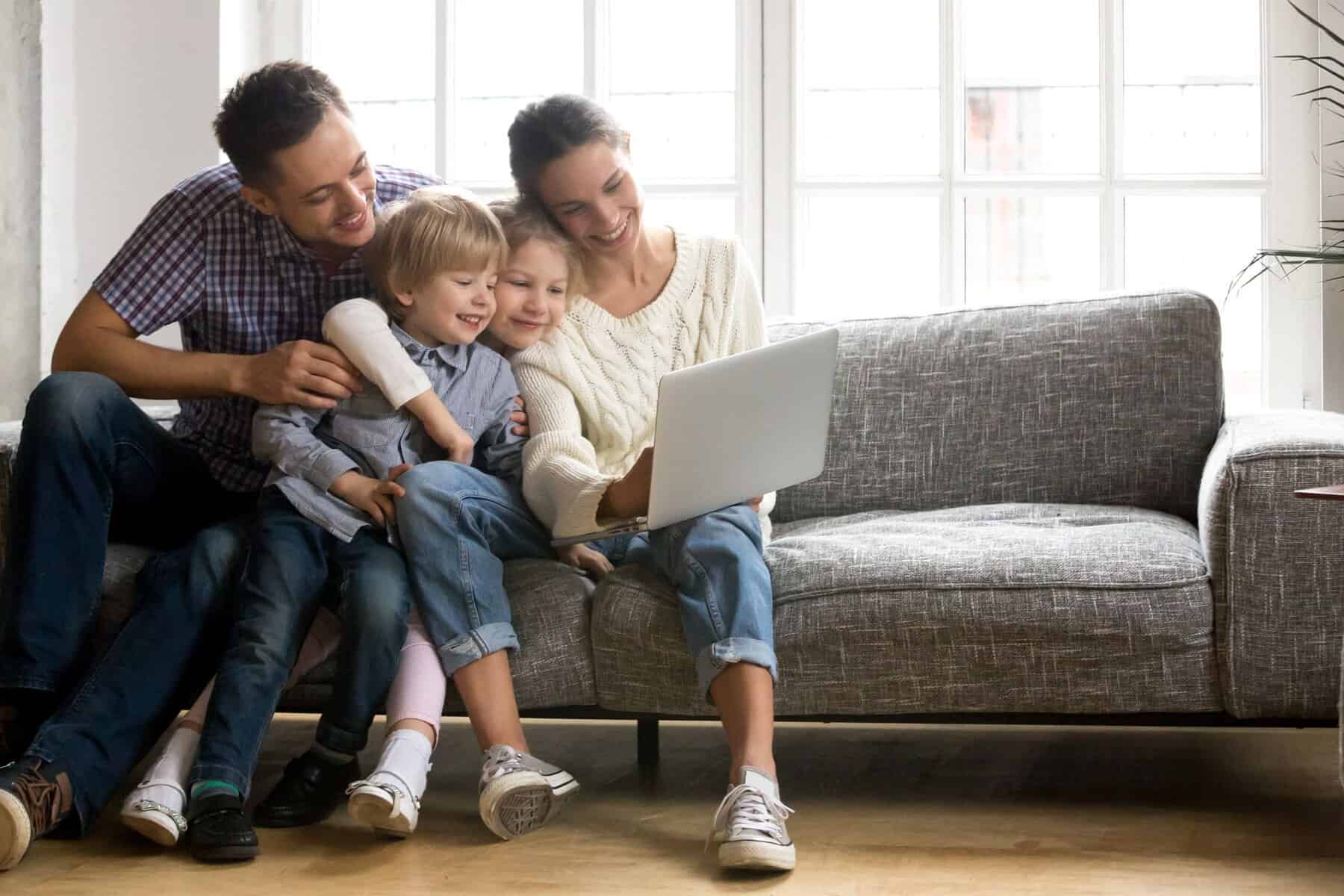 A family of four sits closely together on a gray couch, smiling and looking at a laptop in a bright living room as they research ways to prepare their home for winter and ensure comfort during colder weather.