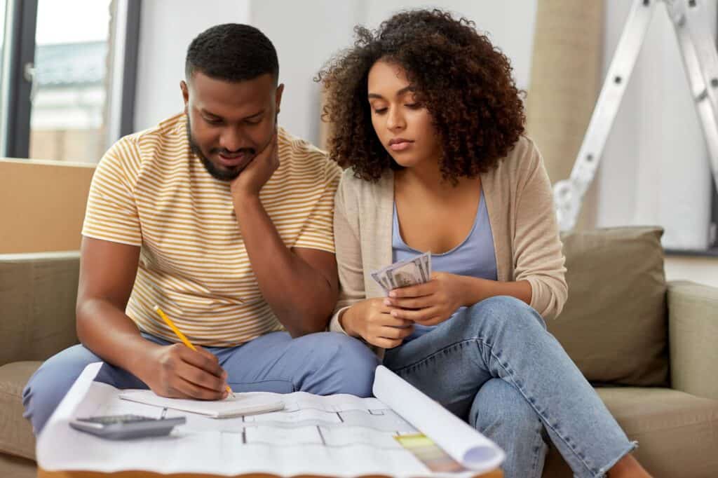 A man writes on paper while a woman holds cash; both sit on a couch with home plans, a calculator, and documents—reviewing options for a new DIY HVAC system.