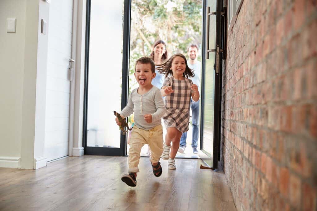 Two young children run excitedly into a home while two adults, likely their parents, smile in the doorway behind them—ready to keep their home warm and save money on heating this season.