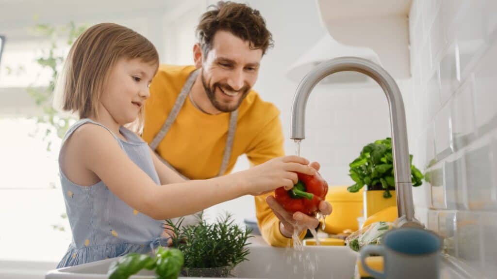 A man and a young girl wash a red bell pepper under a kitchen faucet, surrounded by fresh vegetables and herbs, while discussing kitchen appliance care and the importance of proper garbage disposal maintenance.