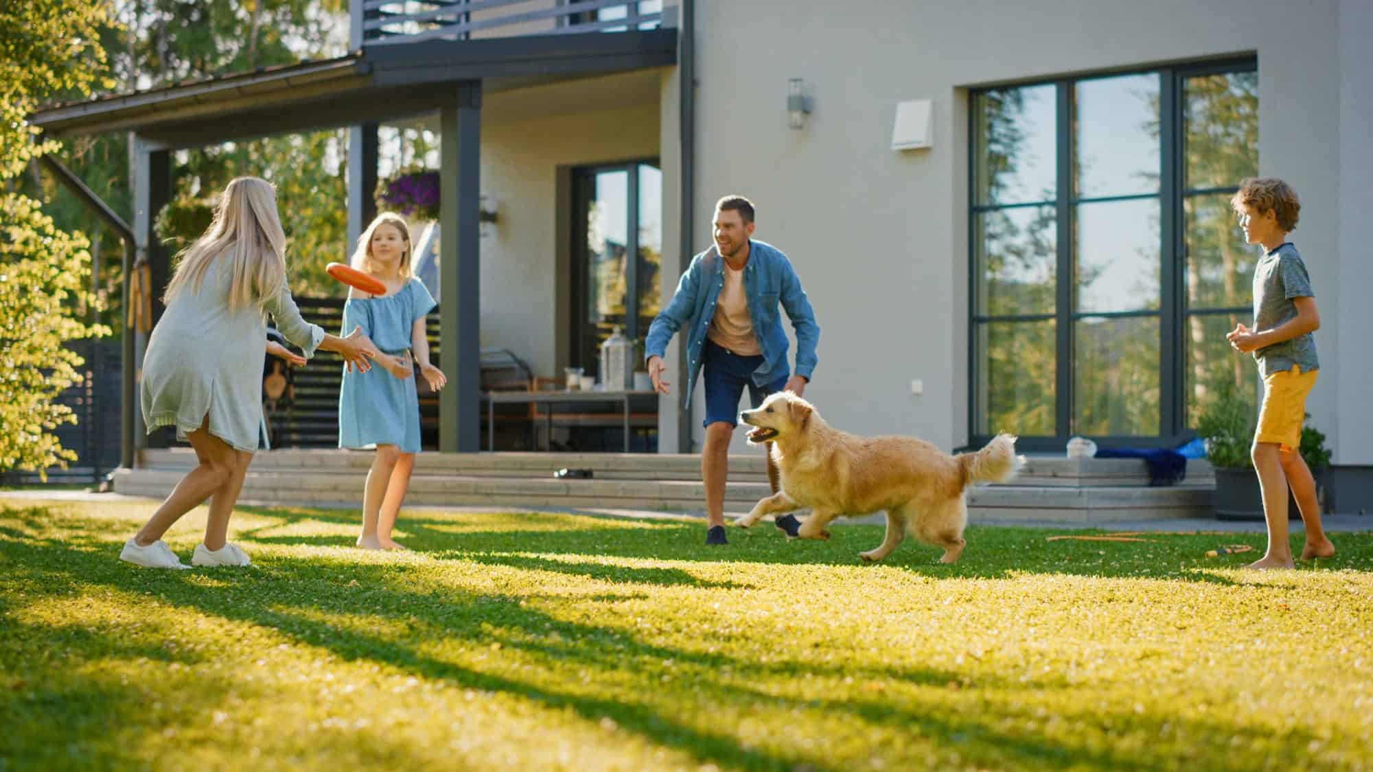 A family with two children and a dog play with a frisbee on the lawn in front of a modern house, where an advanced HVAC system helps alleviate seasonal allergies on this sunny day.