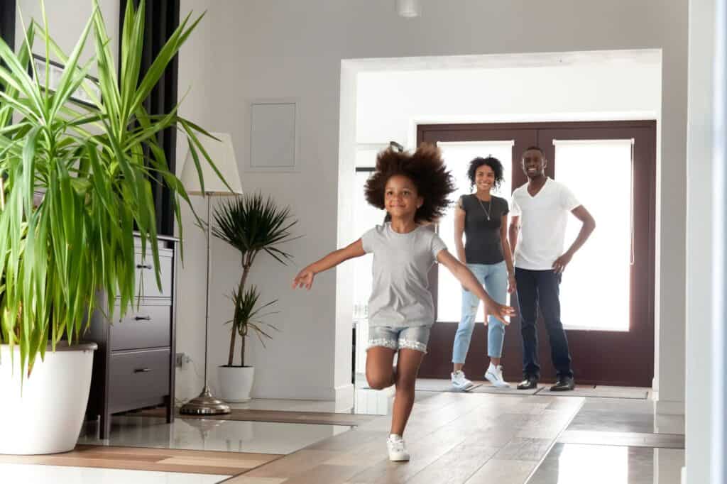 A young girl runs indoors toward the camera while two adults stand together by the doorway in a modern, well-lit home, enjoying comfort thanks to a reliable air conditioning system.