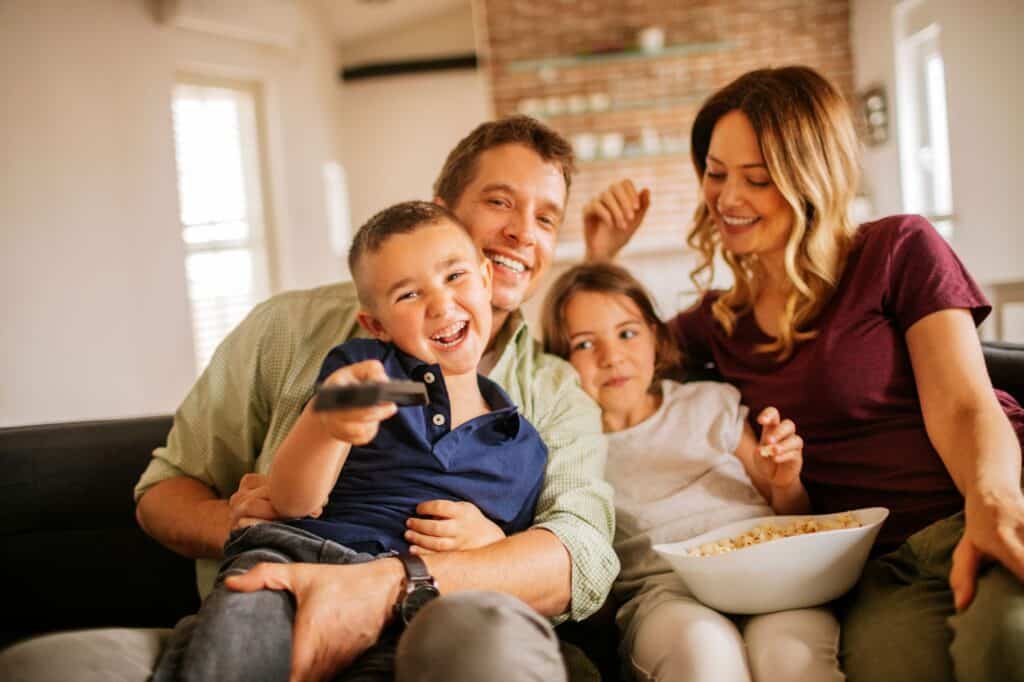 A family of four sits on a couch, smiling and enjoying time together in the comfort provided by a high-efficiency HVAC system. The boy holds a TV remote, the girl holds a popcorn bowl, and the parents sit close to their children.