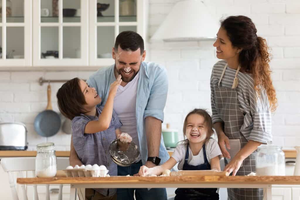 A family of four is gathered in a kitchen, smiling and laughing while baking together, unaware of a pinhole leak in their copper pipes that might need repair or replace options soon—flour and ingredients are scattered across the table.