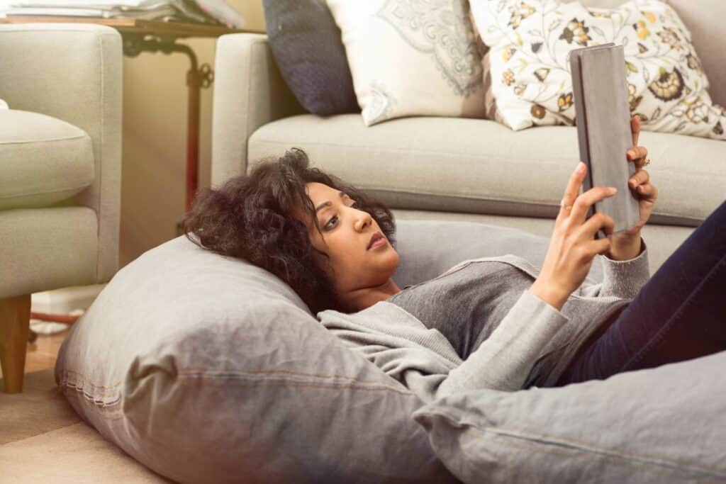 A woman lies on a gray bean bag, holding and looking at a tablet in a living room with sofas and decorative pillows—perhaps researching AC system upgrade options or key AC system considerations for her home.