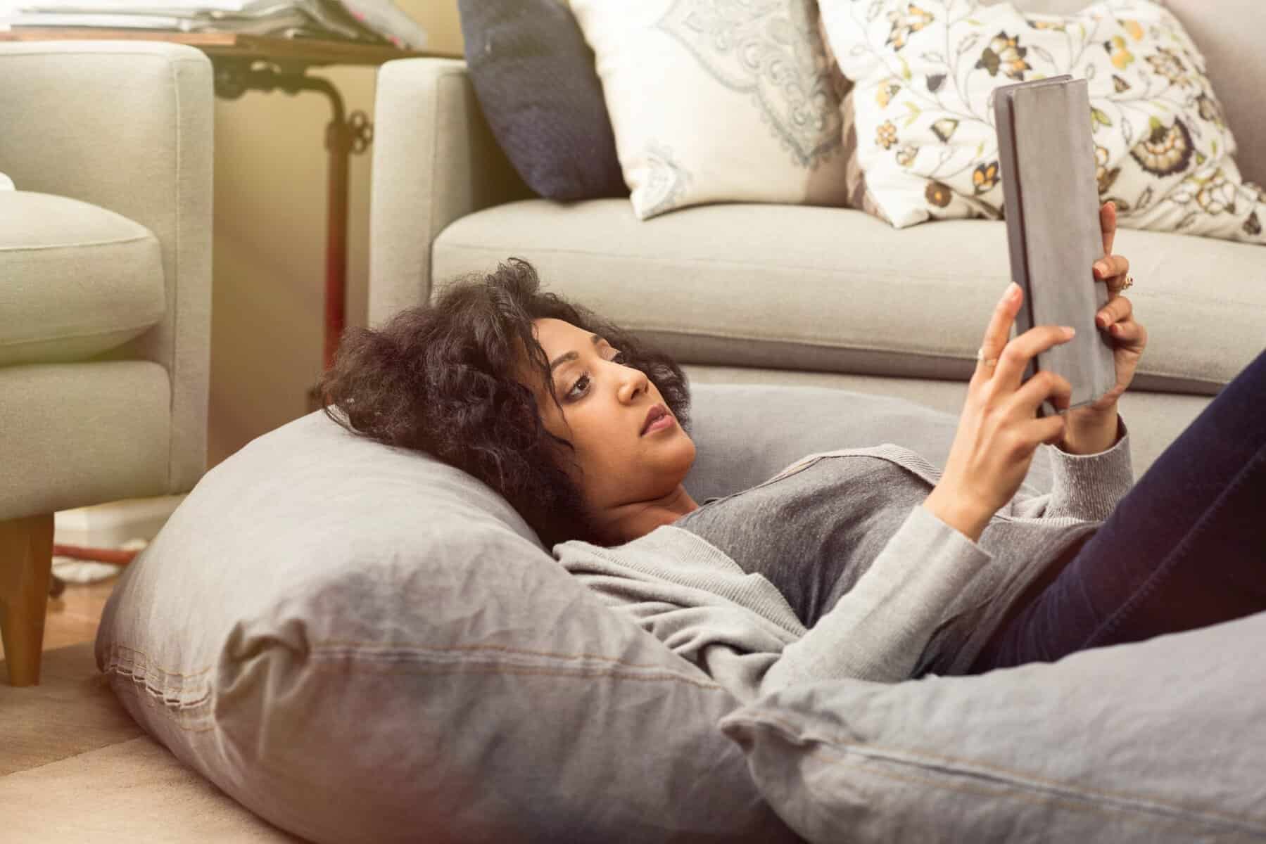 A woman lies on a gray bean bag, holding and looking at a tablet in a living room with sofas and decorative pillows—perhaps researching AC system upgrade options or key AC system considerations for her home.