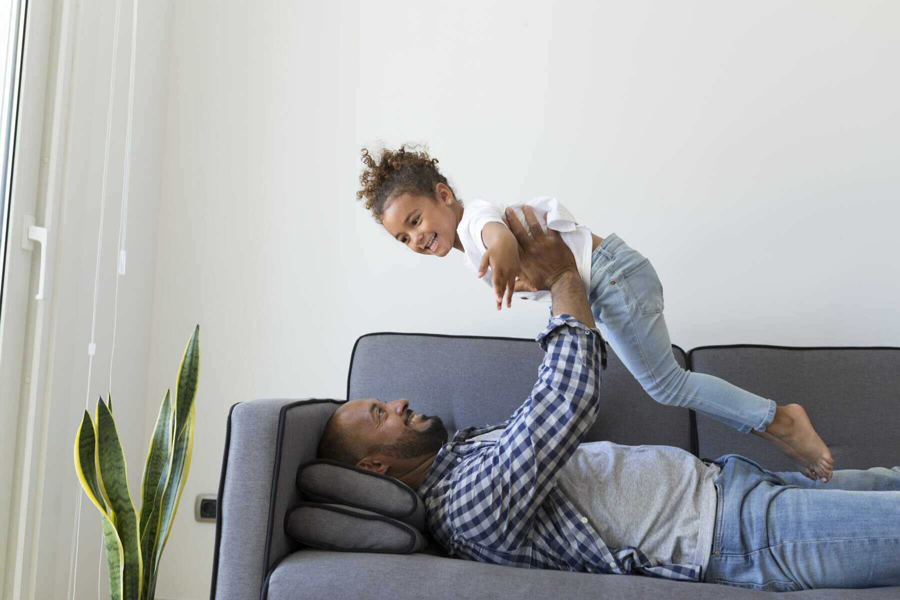 An adult lying on a couch lifts a smiling child into the air; both appear relaxed in a bright living room with a plant nearby, enjoying comfort from an efficient home heating and cooling system.