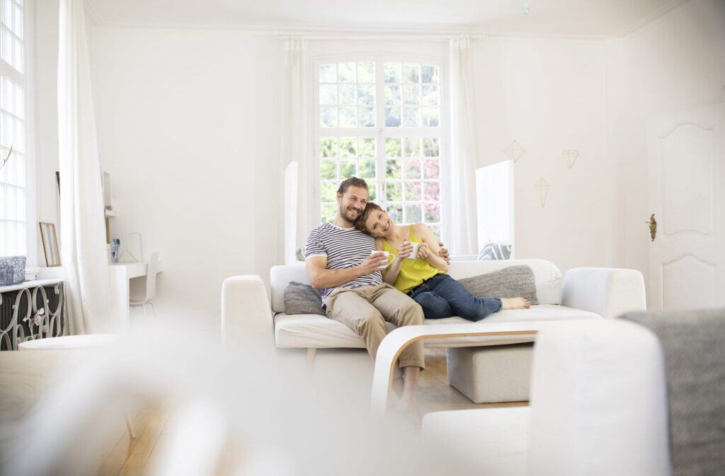 A man and a woman sit closely together on a white sofa in a bright living room, smiling and holding mugs, enjoying the warmth after following DIY Heating System Maintenance tips, with large windows in the background.