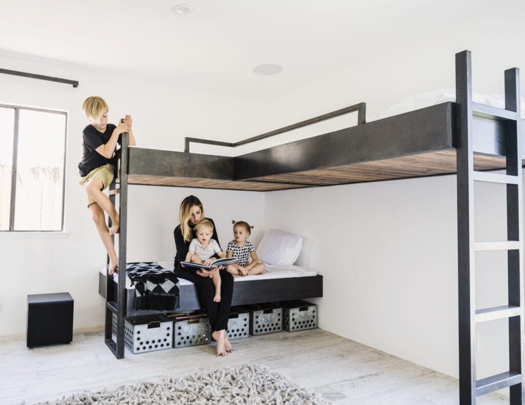 A woman sits on a lower bunk bed with two young children reading a book, while an older child climbs to the upper bunk in a modern, minimal bedroom designed for healthier air and better indoor air quality.