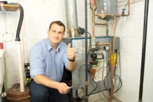 A man in a blue shirt kneels next to a boiler, giving a thumbs up inside a utility room, showcasing excellent customer service.