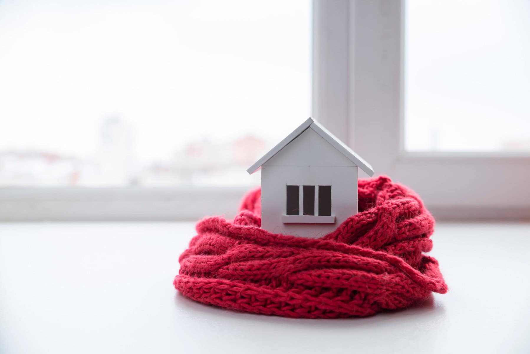 A small model house wrapped in a red knitted scarf sits on a windowsill, symbolizing energy efficient ways to keep home warm, with a blurred cityscape visible outside.
