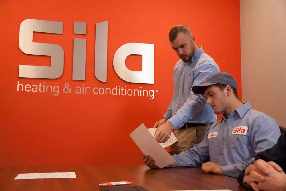 Two Sila employees review a document at a table in front of an orange wall with the Sila heating & air conditioning logo, highlighting the teamwork and dedication you'll find when you learn more about us.