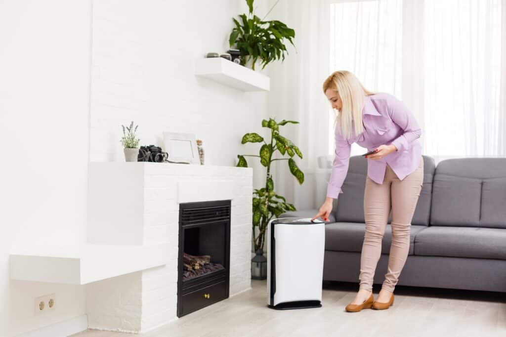 A woman stands in a living room holding a phone and pressing a button on a white air purifier near a gray sofa and green plants—one of the easy ways to control humidity for a comfortable, fresh home environment.