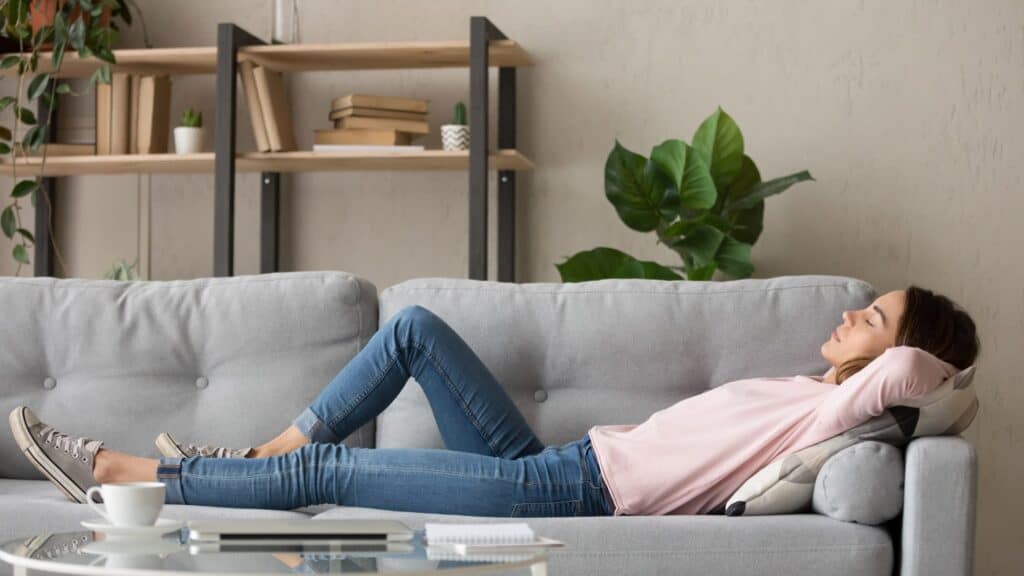 A woman in casual clothes lies on a grey sofa with her hands behind her head, eyes closed, and legs stretched out—enjoying the improved home air quality from effective ways like adding plants seen on shelves in the background.