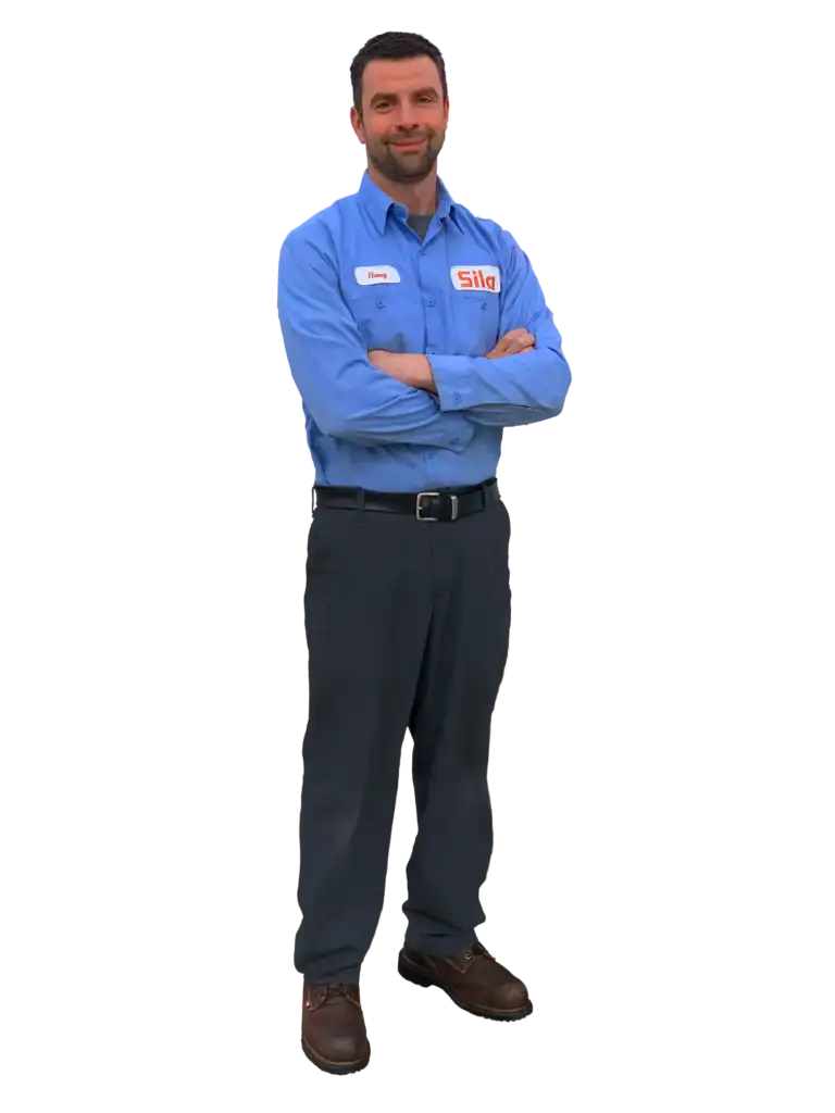 A man wearing a blue uniform shirt with a name tag and the Sila logo, dark pants, and brown shoes stands with arms crossed, facing forward.