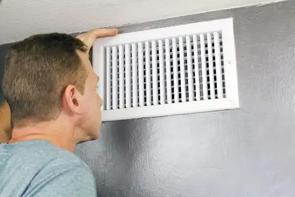 A man in White Plains, NY, closely inspecting a white air vent mounted on a gray wall as part of an air duct installation.