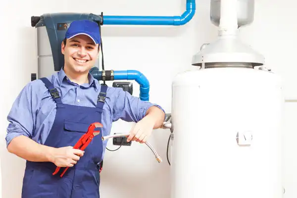 A plumber in blue overalls and cap holds a pipe wrench and flexible hose, standing next to a water heater and pipes—ready for boiler maintenance in White Plains, NY.