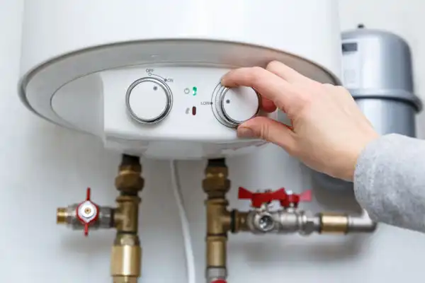 A person adjusts the temperature dial on a white water heater mounted on a wall, part of a recent boiler installation in White Plains, NY, with visible pipes and valves below.