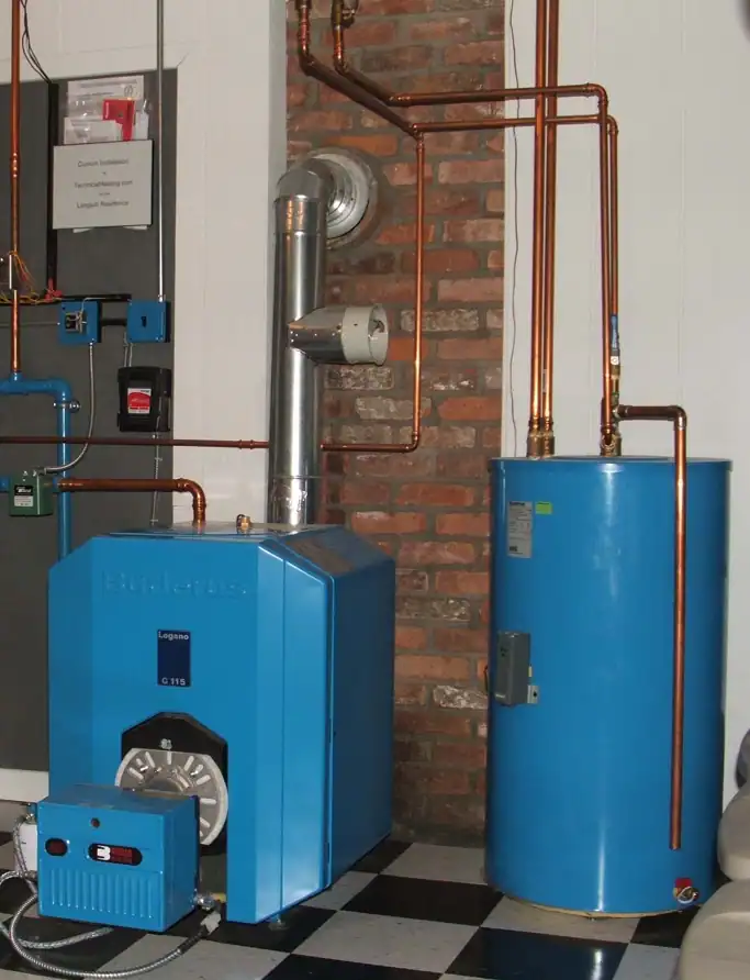 A blue boiler system and hot water tank with connected copper piping installed against a brick and white wall, on a black and white checkered floor.
