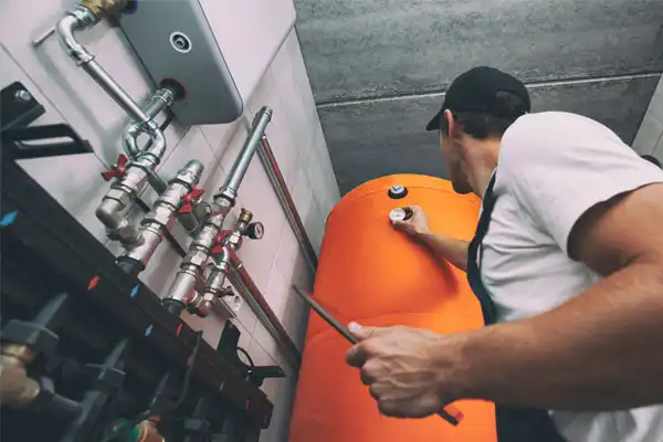 A worker in a cap performs heating repair on an orange industrial water heater, adjusting a valve with a tool; visible pipes and gauges are mounted on the wall nearby.