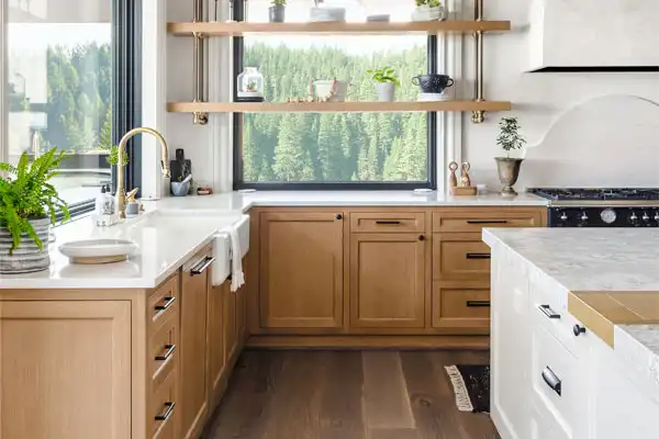 Modern kitchen in White Plains, NY, with light wood cabinets, white countertops, open shelves holding plants, a black stove, and a large window overlooking forest scenery. Expert plumbing repair available to keep everything running smoothly.
