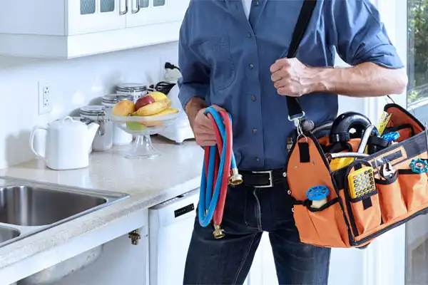 A plumber in a blue shirt carries a tool bag and hoses, standing in a bright White Plains NY kitchen near a sink and countertop with fruit and containers, ready for kitchen plumbing repair.