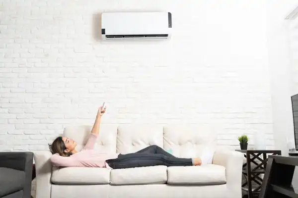 A person lies on a sofa holding a remote control, pointing it at a wall-mounted air conditioner after a recent mini-split installation in their bright, modern White Plains NY living room.