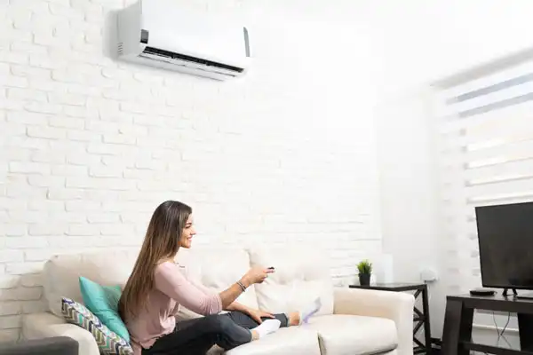 A woman sits on a couch in a living room in White Plains, NY, using a remote control to operate a wall-mounted air conditioner, emphasizing the importance of regular mini-split maintenance for optimal comfort.