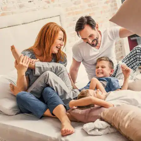 A family of four laughs and plays together on a bed, with parents and two children engaged in a pillow fight, enjoying the comfort brought by recent High Velocity Air Conditioning Repair.