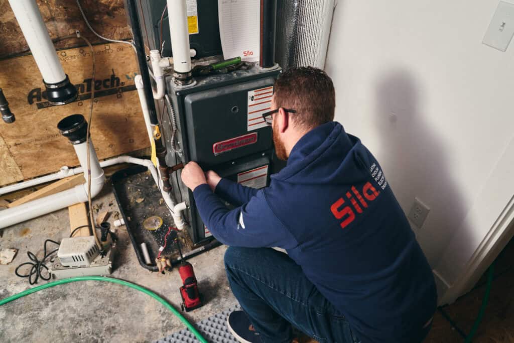 A technician in a blue hoodie kneels and works on a furnace in a utility room with exposed pipes and tools nearby.