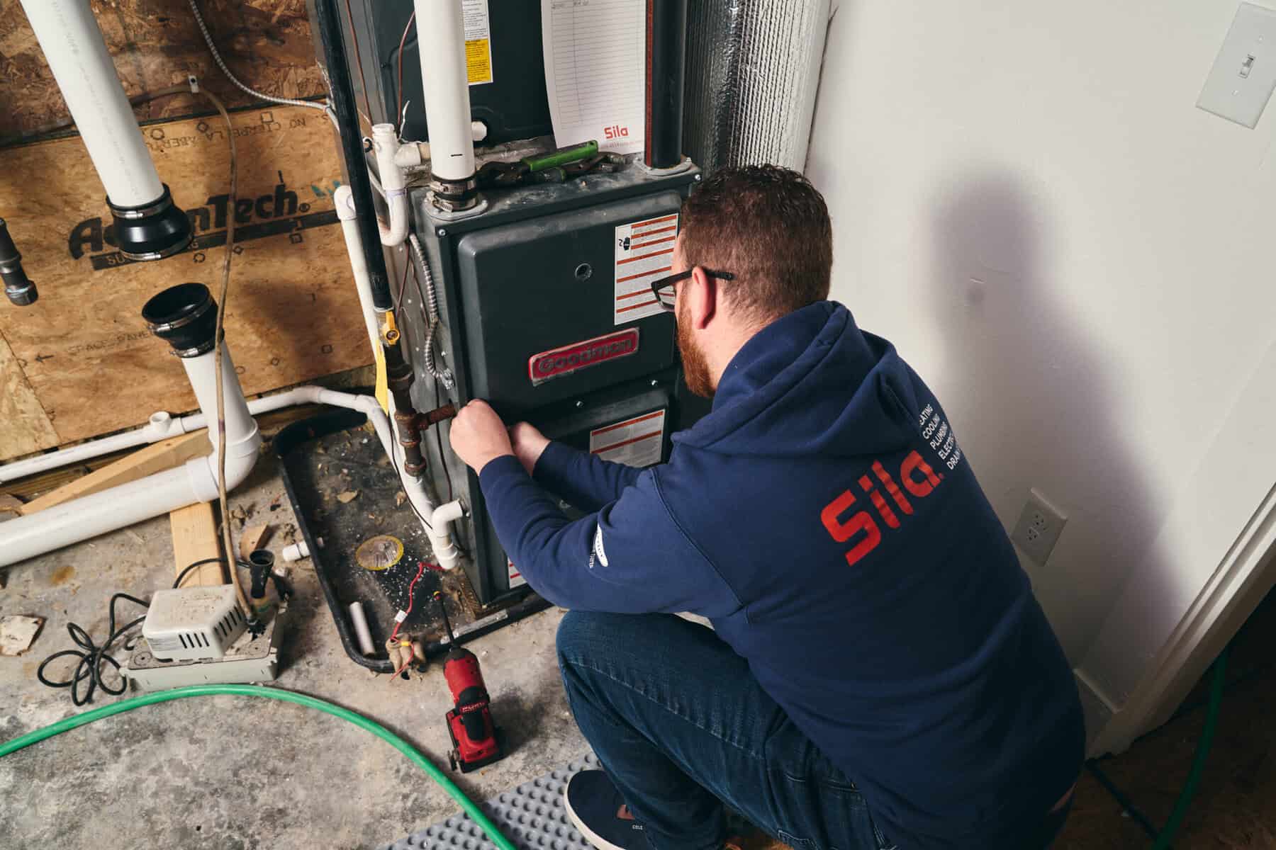 A technician in a blue hoodie kneels and works on a furnace in a utility room with exposed pipes and tools nearby.